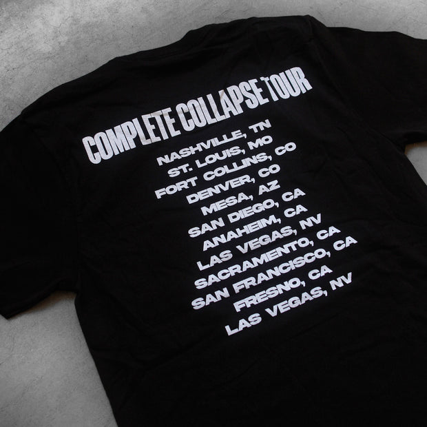 close up, angled image of the back of a black tee shirt laid flat on a concrete floor. tee has a full back print in white. at the top says complete collapse tour, with the locations listed below.