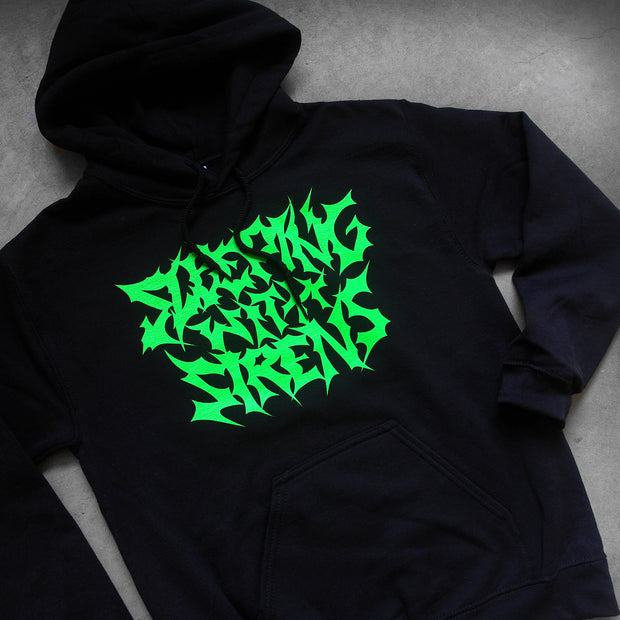 close up, angled image of a black pullover hoodie laid flat on a concrete floor. front of the hoodie has green print in metal font that says sleeping with sirens