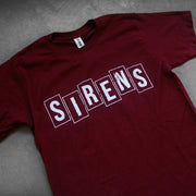 close up, angled image of a maroon tee shirt laid flat on a concrete floor. the front of the shirt has a center chest print in white that says sirens.
