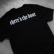 close up, angled image of the back of a black tee shirt laid flat on a concrete floor. back of the tee has a white print at the top across the shoulders that says there's the door